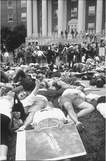 Black and white photo of thousands of demonstrators lying down on the lawn, surrounded by police on horseback, in front of a building with pillars.