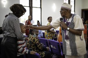 A reverend holds a mic and holds out his hand to a child and his grandmother in a church.