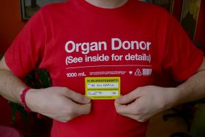 Close up of someone's chest, wearing a red shirt that says "Organ Donor" and holding yellow card identifying them as an organ donor.