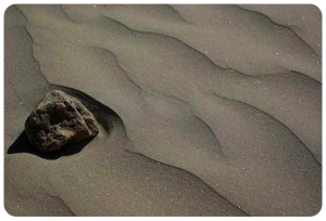 A craggy-textured rock is on the rippled sandy shore of a beach.