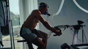 An older male on a stationary bike is breathing into a tube in order to measure his breath.