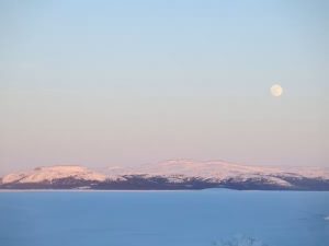 Snowcapped mountain range with snow in front and a full moon rising above it.