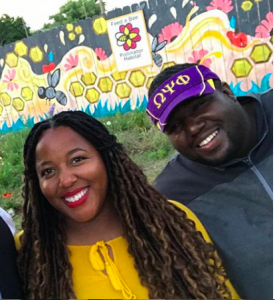 Nicole Lindsey in yellow and Timothy Jackson in a purple and yellow visor smile in front of a bee mural painted on a fence