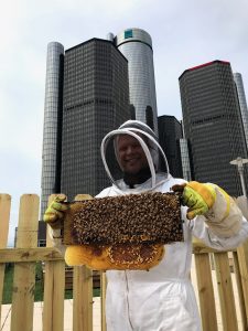 Brian Peterson-Roest in a full bee suit holds up a frame of honey bees in front of tall building in background