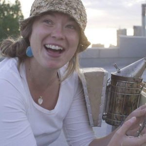 Eliese Watson in white and a straw hat sits outside by a bee smoker