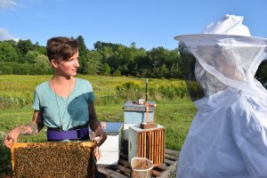 Ang Roell holding beehive frame and looking at someone in full bee suit