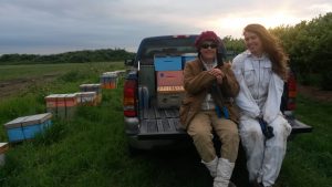 Julia Common in a red hat & Sarah Common in a bee suit sit in back a pickup truck in a field with beehives to their left