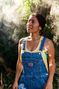 Jasmine Joy in yellow tank top and overalls looks left toward smoke from bee smoker