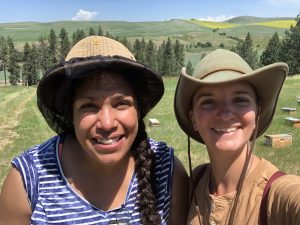 New Mexico-based beekeeper Melanie Kirby and Ang Roell smile at the camera in a field with beehives in the background