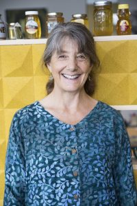 Marla Spivak smiles at camera standing in front of yellow background with honey jars on ledge