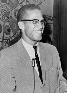 Black-and-white photograph of Malcolm X turned 45 degrees laughing at something out of sight. He is wearing a suit and in front of a microphone.