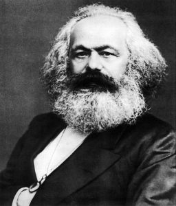 Black and white portrait of Karl Marx's head and upper body. Marx looks solemnly at the camera with his hand tucked into his vest.
