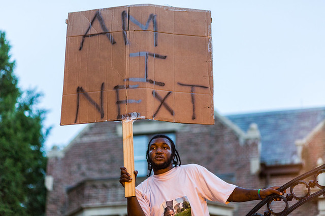 Photo of a young black man from the chest up pictured holding a cardboard sign with "Am I Next" spray-painted on it.