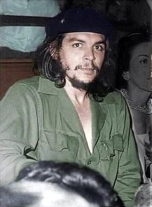 Che Guevara sitting amongst people barely visible wearing iconic green military suit and beret, looking directly into camera.