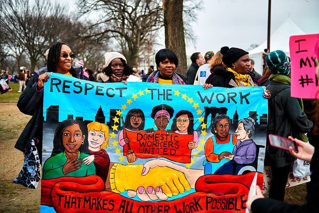 Four black women holding up a colorful banner that read "respect the work that makes all other work possible" with images of different domestic work elements. Pictured at the Women's March, 2017.