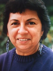 Photograph of Anzaldúa's head and shoulders. She is wearing a blue sweater and beaded earrings, while smiling and looking at the camera. The backdrop is out-of-focus greenery