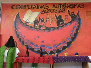 Mural on a wall that says Cooperativas Autonomas Zapatistas. The mural consists of members of the EZLN with ski masks in a boat-like structure with unmasked women and children