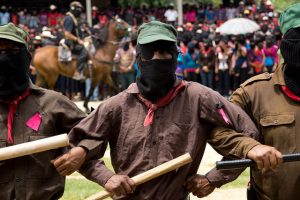 Three Zapatista men in uniform linking arms with bats in their hands. There is a crowd of other zapatistas in the background in what looks like a ceremony with a horse.