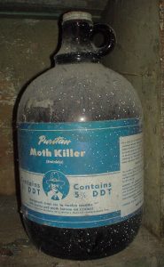 Large glass jug with white paint dots in a basement that has a blue label that says, "Moth Killer" and advertises that the solution contains "5% DDT"